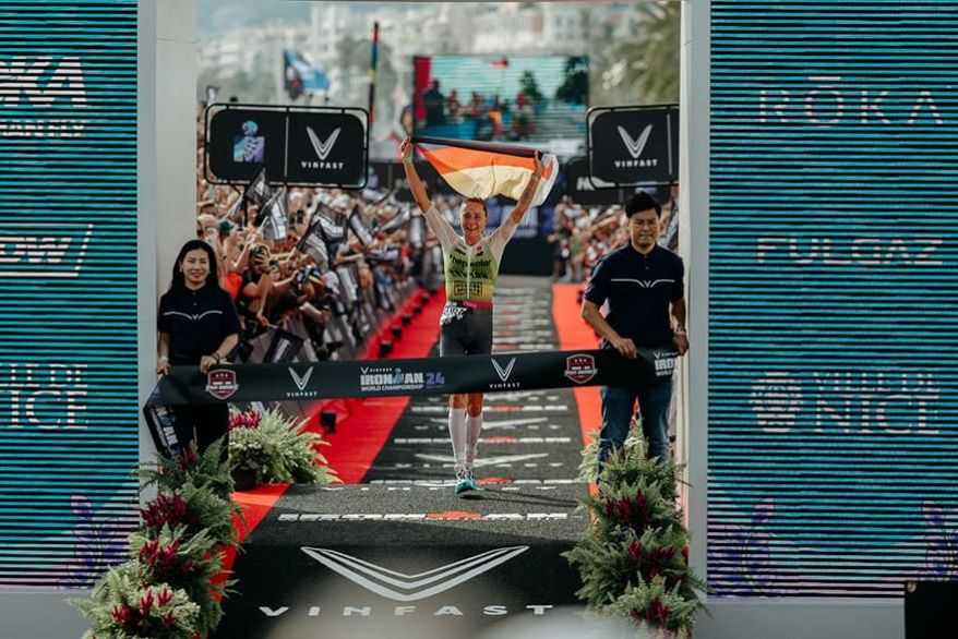 Das Bild zeigt Laura Philipp, wie sie die Ziellinie der Ironman-Weltmeisterschaft 2024 überquert und eine deutsche Flagge über ihrem Kopf hält. Sie lächelt und geht unter dem Banner hindurch, auf dem “VinFast Ironman 24 World Championship” steht. Zuschaue